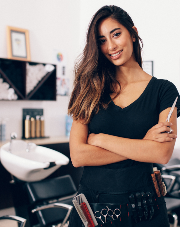 Cosmetology school student with arms crossed smiles wearing tool belt